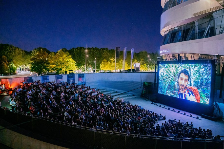 Open-air cinema at night with many viewers in front of a large screen. Trees and a modern building can be seen in the background., © Mercedes-Benz Heritage GmbH Open-air cinema at night with many viewers in front of a large screen. Trees and a modern building can be seen in the background., © Mercedes-Benz Heritage GmbH