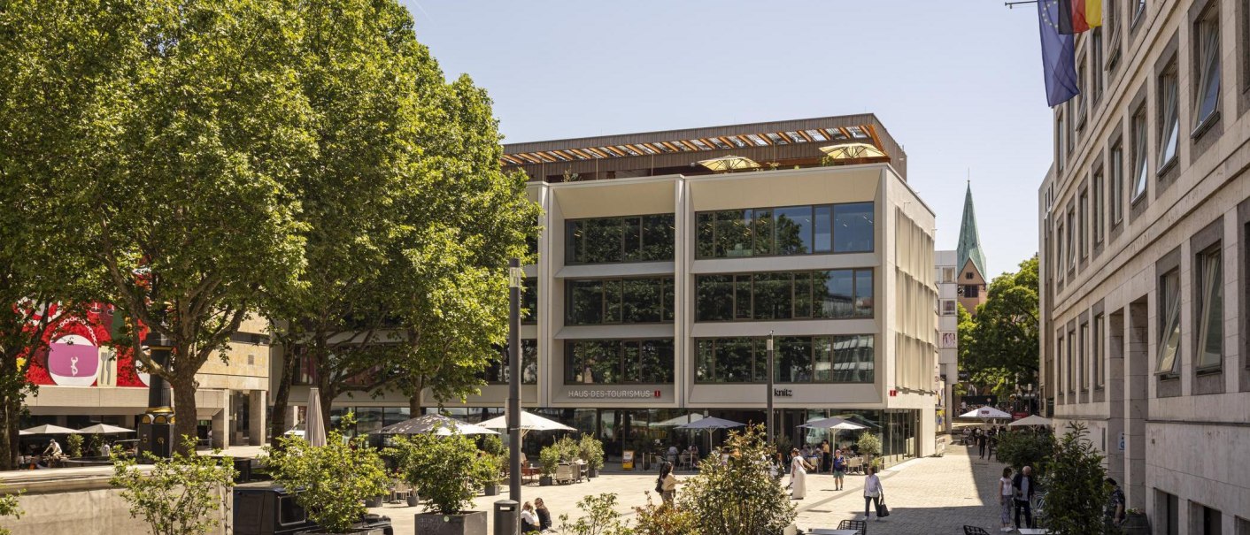 Modernes Gebäude mit Café und Bäumen auf einem sonnigen Platz. Menschen flanieren und genießen die Atmosphäre., © SMG, Sarah Schmid Modernes Gebäude mit Café und Bäumen auf einem sonnigen Platz. Menschen flanieren und genießen die Atmosphäre., © SMG, Sarah Schmid