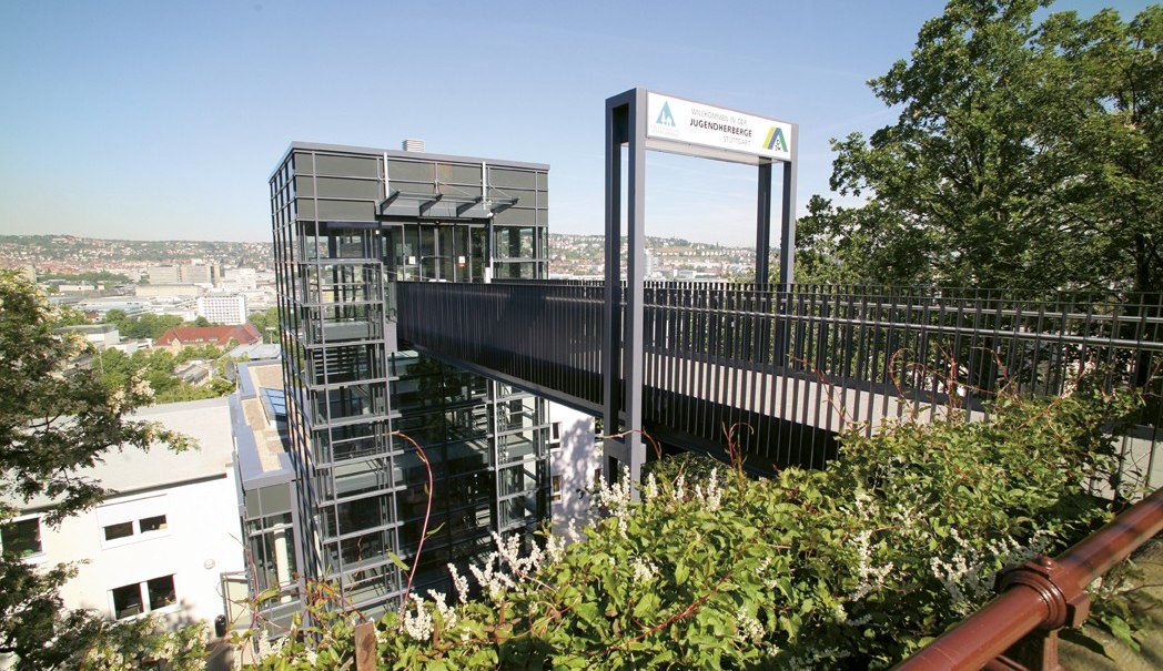 Modern youth hostel in Stuttgart with glass façade and bridge, surrounded by green vegetation and city view in the background., © TOMAS Modern youth hostel in Stuttgart with glass façade and bridge, surrounded by green vegetation and city view in the background., © TOMAS