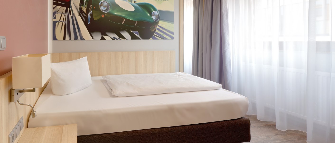 Modernes Einzelzimmer mit Bett, großem Fenster und Vorhängen. An der Wand ein Bild von Autos., © attimo Hotel Stuttgart Modernes Einzelzimmer mit Bett, großem Fenster und Vorhängen. An der Wand ein Bild von Autos., © attimo Hotel Stuttgart