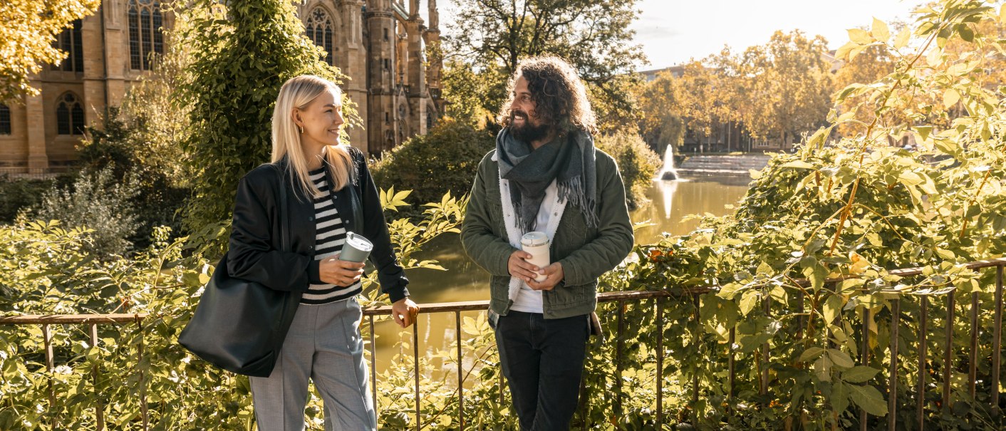 Zwei Personen stehen lächelnd mit Kaffeebechern vor einer Kirche und einem See. Die Szene ist herbstlich und sonnig., © SMG Stuttgart Marketing GmbH - Sarah Schmid Zwei Personen stehen lächelnd mit Kaffeebechern vor einer Kirche und einem See. Die Szene ist herbstlich und sonnig., © SMG Stuttgart Marketing GmbH - Sarah Schmid