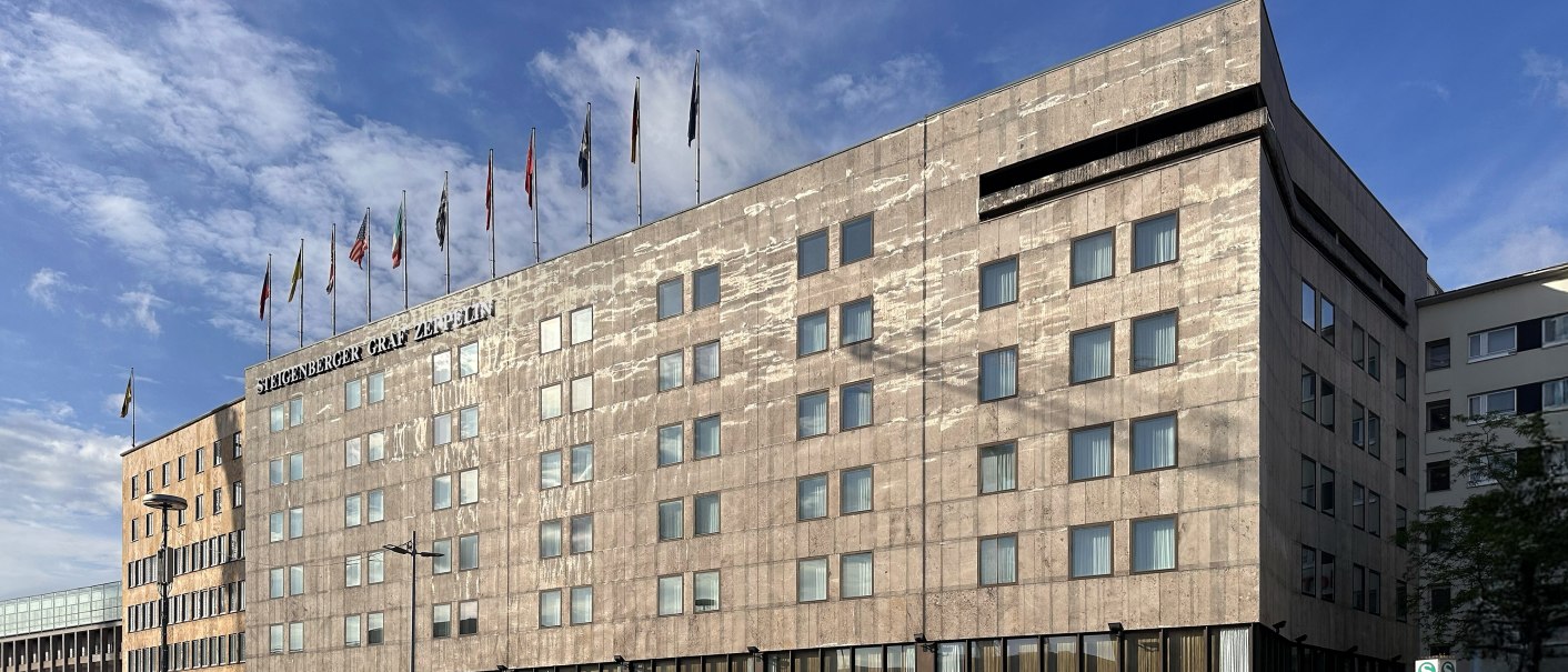 The Steigenberger Graf Zeppelin Hotel with flags on the roof against a blue sky. Modern architecture with many windows., © Steigenberger Graf Zeppelin