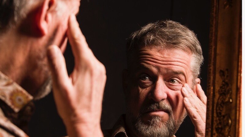 A man with a beard looks thoughtfully into a mirror and touches his face. The background is dark, the frame of the mirror is decorative., &copy; Rosenau Kultur e.V.