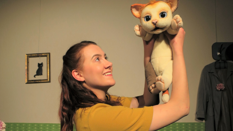 Eine Frau hebt eine Katzenpuppe in die Luft und lächelt. Im Hintergrund hängt ein Bild einer schwarzen Katze an der Wand., © FITHE – Das Figurentheater aus Ostbelgien Eine Frau hebt eine Katzenpuppe in die Luft und lächelt. Im Hintergrund hängt ein Bild einer schwarzen Katze an der Wand., © FITHE – Das Figurentheater aus Ostbelgien