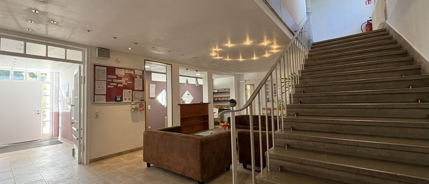 Heller Eingangsbereich mit Treppe, braunen Sofas und Infotafel. Moderne Beleuchtung und Fliesenboden schaffen eine einladende Atmosph&auml;re., &copy; TOMAS