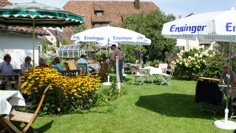 Ein sommerlicher Garten mit Menschen, die unter Sonnenschirmen an Tischen sitzen, umgeben von bl&uuml;henden Blumen und gr&uuml;ner Vegetation., &copy; Weinbau Knobloch-Wolfrum
