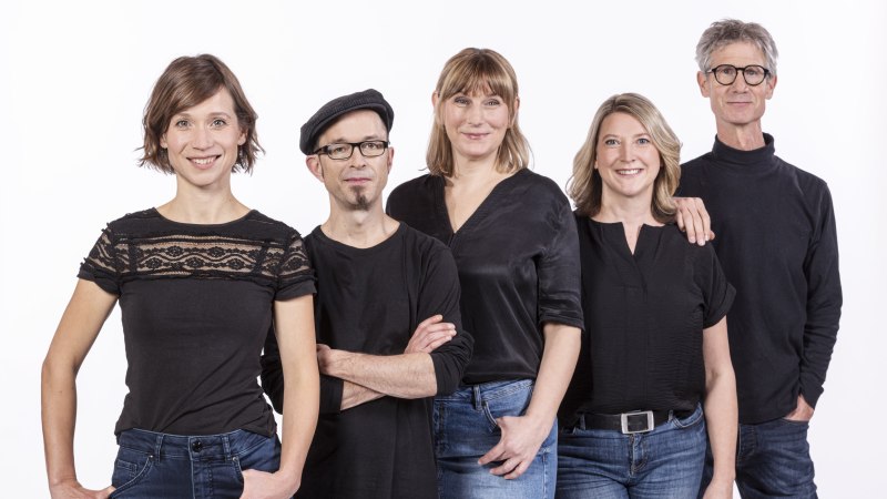 F&uuml;nf Personen in schwarzer Kleidung stehen nebeneinander und l&auml;cheln in die Kamera. Der Hintergrund ist wei&szlig;., &copy; Improtheater Stuttgart / Rainer Wengel Fotografie