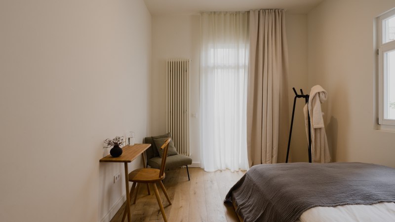 Minimalistisch eingerichtetes Schlafzimmer mit Holzboden, Bett, kleinem Schreibtisch und Stuhl. Vorh&auml;nge und ein Bademantel auf einem St&auml;nder erg&auml;nzen das Bild., &copy; Peter Oliver Wolff / DAS EDITH