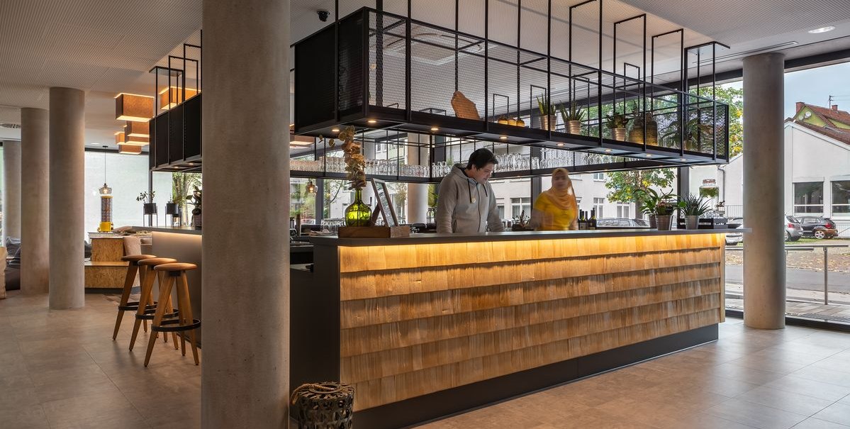 Moderne Rezeption mit Holzverkleidung und Hängeregalen. Zwei Personen stehen hinter dem Tresen. Große Fenster im Hintergrund., © Novum Hospitality