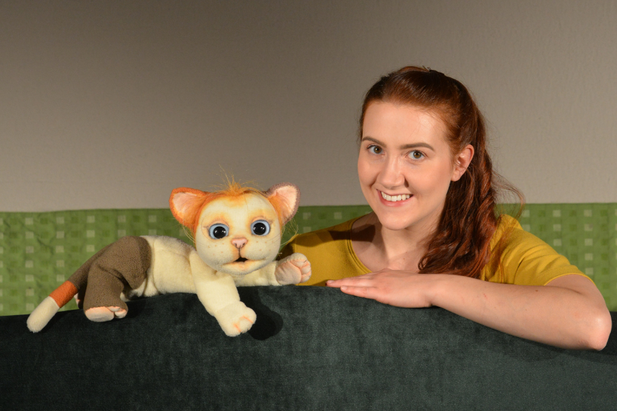 Eine Frau mit braunen Haaren lächelt neben einer niedlichen Katzenpuppe mit großen Augen. Beide sind hinter einem grünen Hintergrund platziert., © FITHE – Das Figurentheater aus Ostbelgien Eine Frau mit braunen Haaren lächelt neben einer niedlichen Katzenpuppe mit großen Augen. Beide sind hinter einem grünen Hintergrund platziert., © FITHE – Das Figurentheater aus Ostbelgien