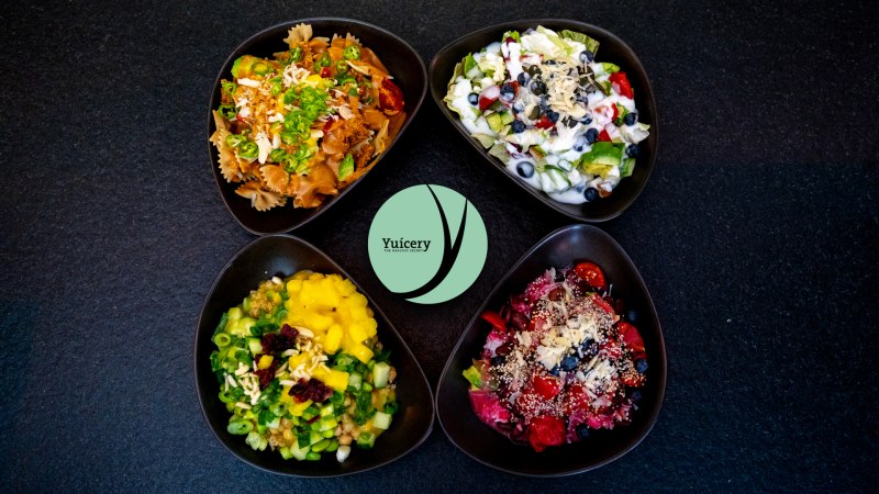 Four bowls with colorful salads and ingredients, including pasta, fruit and vegetables, on a black background. Yucery" logo in the center., © Yuicery Four bowls with colorful salads and ingredients, including pasta, fruit and vegetables, on a black background. Yucery" logo in the center., © Yuicery