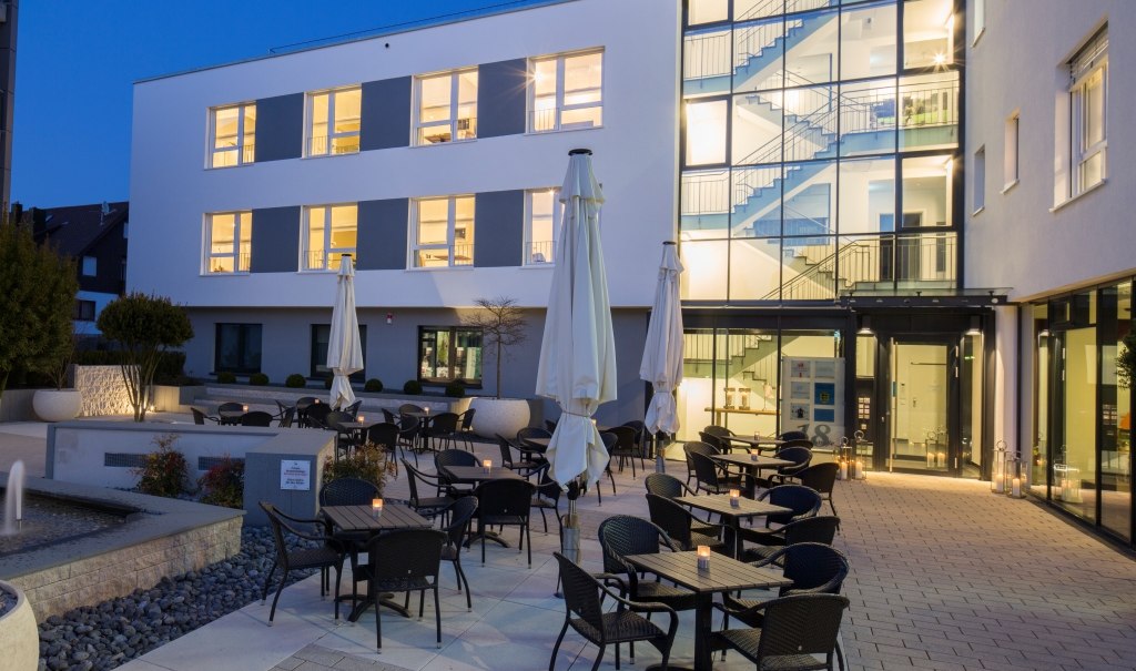 Modern hotel exterior at dusk with illuminated windows, terrace with tables and chairs and large parasols., © Hotel LOGIS Modern hotel exterior at dusk with illuminated windows, terrace with tables and chairs and large parasols., © Hotel LOGIS