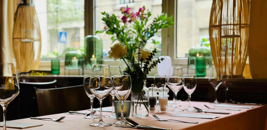 A stylishly laid table in a café with wine glasses, cutlery and flower arrangements in front of a window., © FreCla Gastronomiebetriebe GmbH