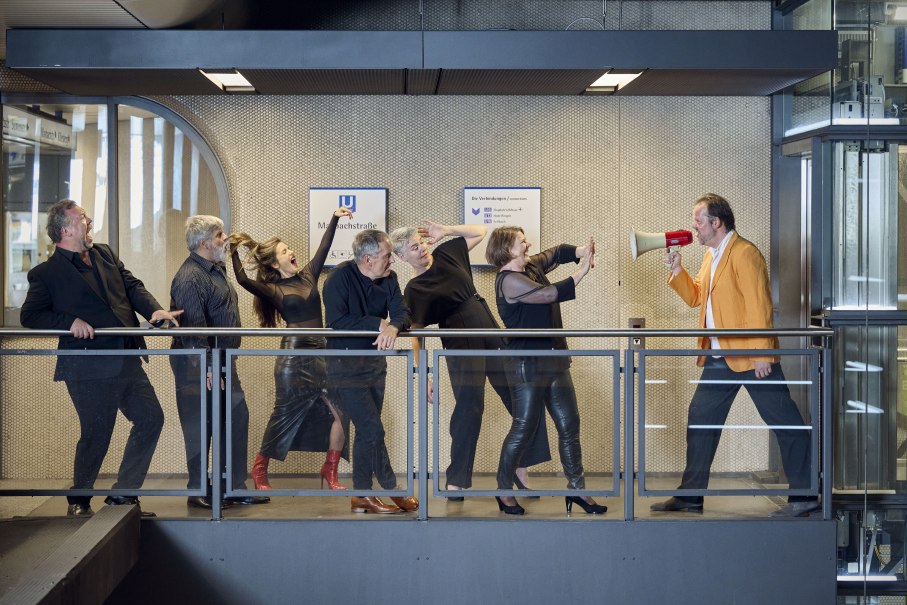 Eine Gruppe von Menschen steht auf einem Balkon. Eine Person im orangefarbenen Sakko spricht in ein Megafon, während die anderen reagieren., © Martin Sigmund Eine Gruppe von Menschen steht auf einem Balkon. Eine Person im orangefarbenen Sakko spricht in ein Megafon, während die anderen reagieren., © Martin Sigmund