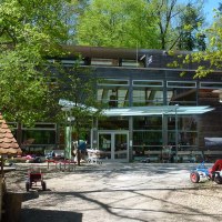 Inner courtyard of the House of the Forest with toy vehicles, trees and a small wooden house. Modern glass façade in the background., © Haus des Waldes