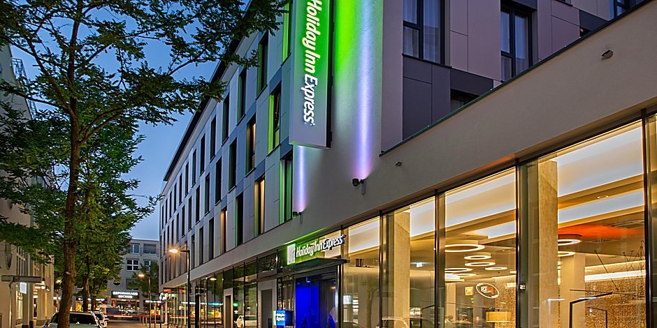 Außenansicht eines modernen Holiday Inn Express Hotels bei Dämmerung, mit beleuchtetem Logo und Glasfassade., © Holiday Inn Express Waiblingen Außenansicht eines modernen Holiday Inn Express Hotels bei Dämmerung, mit beleuchtetem Logo und Glasfassade., © Holiday Inn Express Waiblingen