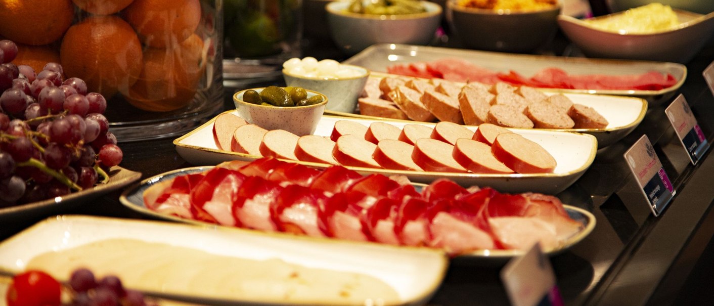 A breakfast buffet with various types of sausage, grapes, oranges and pickled cucumbers arranged on plates and bowls., &copy; MASEVEN Stuttgart SI-Centrum