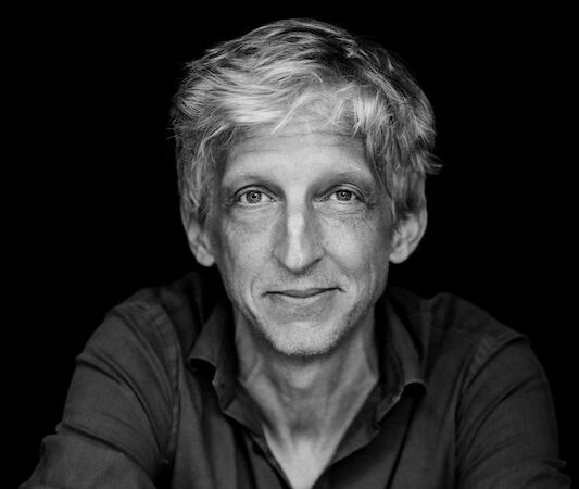Black and white portrait of a man with light-colored hair, smiling and looking into the camera. He has his arms crossed and is sitting in front of a dark background., &copy; Renitenztheater