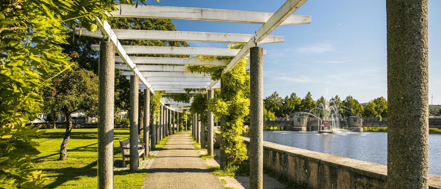 A sunny park path with a pergola and green plants leads along a lake with a fountain in the background., © SMG Stuttgart Marketing GmbH - Sarah Schmid A sunny park path with a pergola and green plants leads along a lake with a fountain in the background., © SMG Stuttgart Marketing GmbH - Sarah Schmid