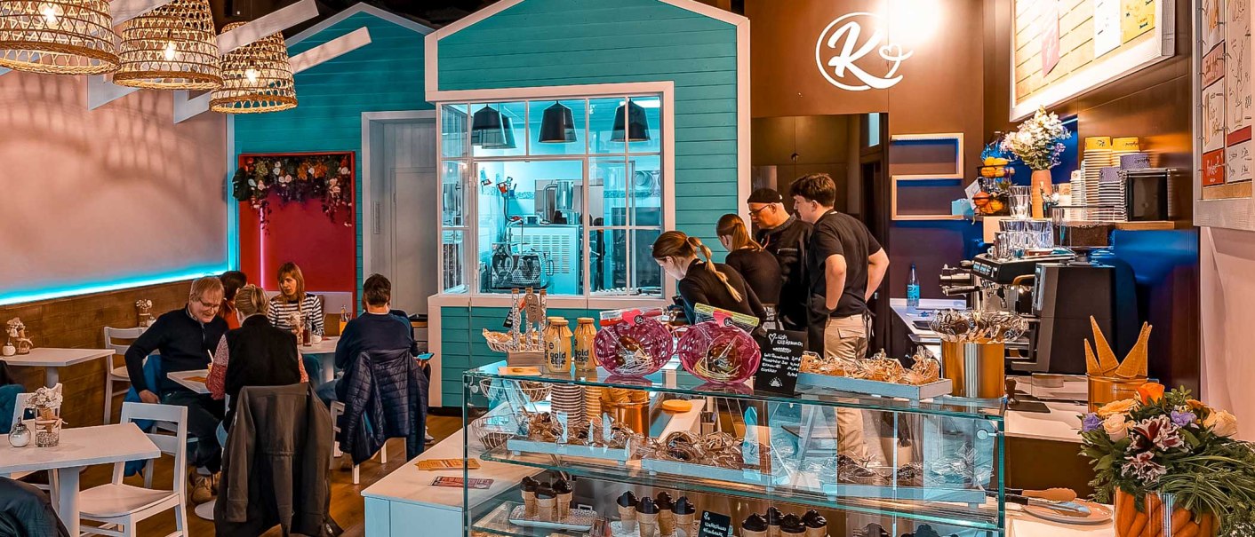 Gemütliches Café mit Gästen an Tischen, Personal hinter der Theke. Dekorative Beleuchtung und bunte Einrichtung schaffen eine einladende Atmosphäre., © Kesselglück Gemütliches Café mit Gästen an Tischen, Personal hinter der Theke. Dekorative Beleuchtung und bunte Einrichtung schaffen eine einladende Atmosphäre., © Kesselglück