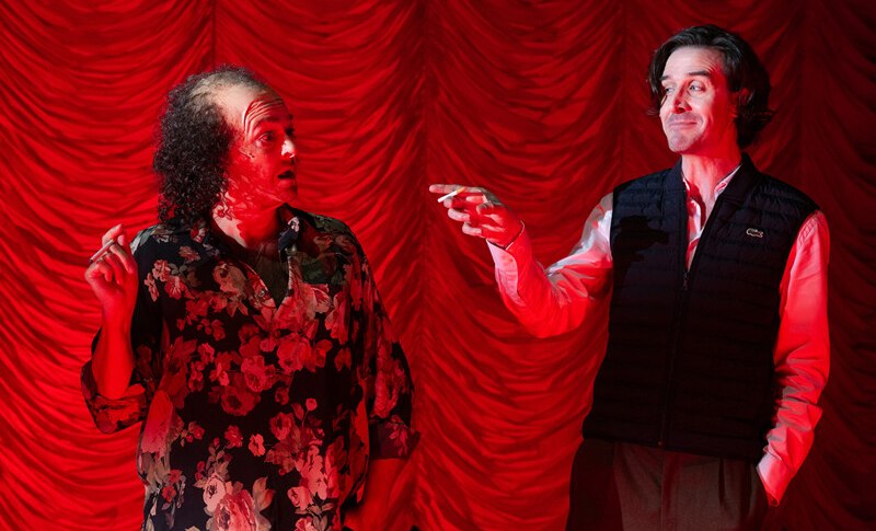 Zwei M&auml;nner stehen vor einem roten Vorhang. Der linke tr&auml;gt ein Blumenhemd, der rechte eine Weste. Beide gestikulieren lebhaft., &copy; W&uuml;rttembergische Staatstheater Stuttgart