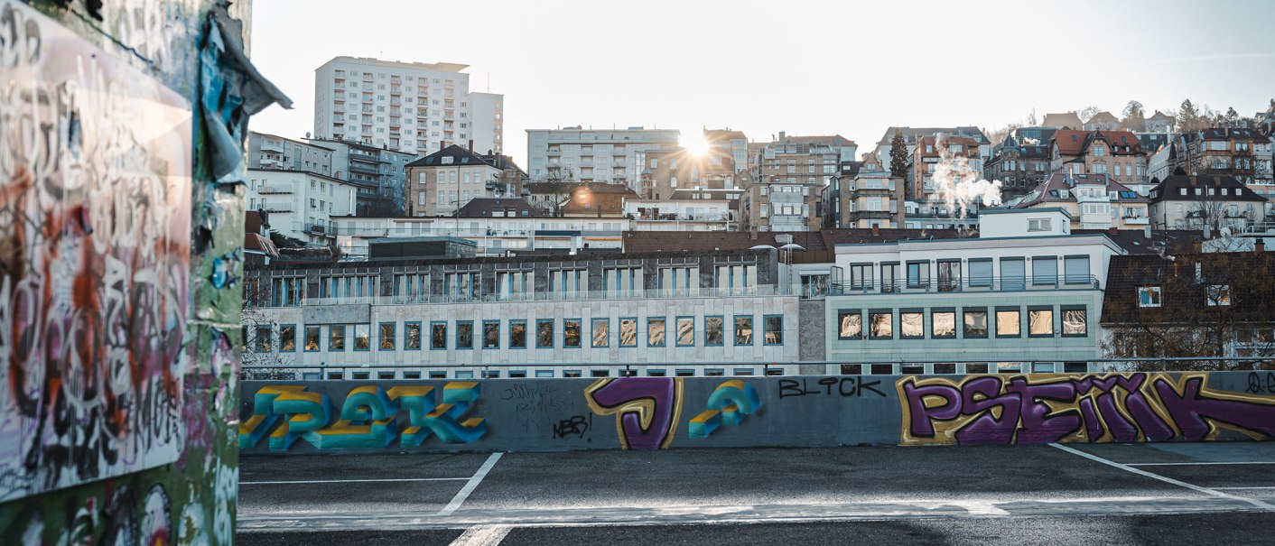 Städtische Skyline von Stuttgart bei Sonnenuntergang, mit Graffiti auf einer Mauer im Vordergrund. Gebäude und Rauch steigen in den Himmel., © SMG Stuttgart Marketing GmbH -Sarah Schmid