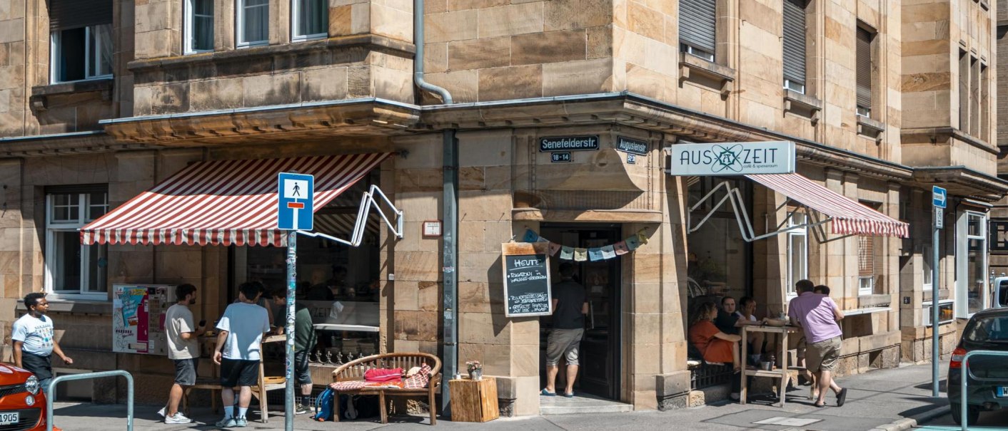 Cafébar Auszeit an einer Straßenecke mit roter Markise. Menschen sitzen draußen, Straßenschilder und Autos sind sichtbar., © SMG, Sarah Schmid