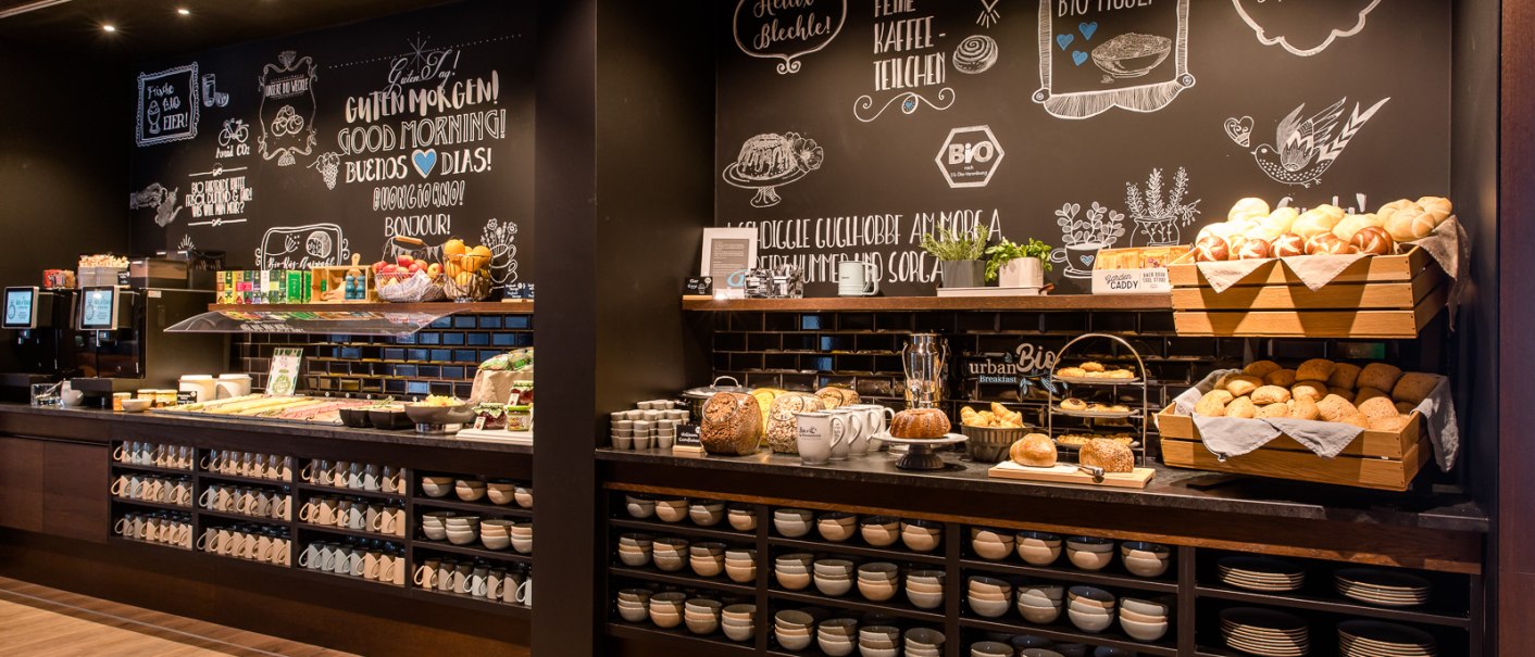 Ein Frühstücksbuffet mit Brotkörben, Tassen und Schüsseln vor einer dekorativen Kreidetafelwand., © TOMAS