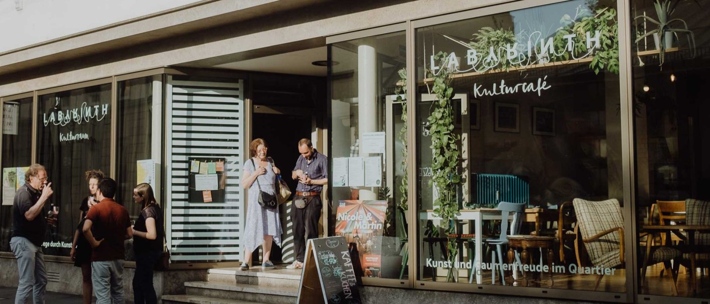 People chat in front of the LABYRINTH cultural center & café. The windows are decorated with plants and there is a sign in front of the entrance., © Christoph Steinweg