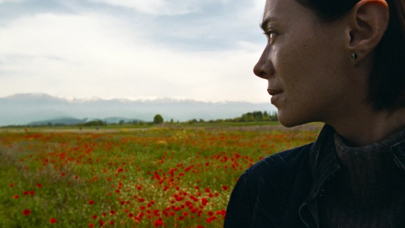 Eine Frau blickt nachdenklich über ein weites Mohnblumenfeld mit Bergen im Hintergrund., © MUBI Eine Frau blickt nachdenklich über ein weites Mohnblumenfeld mit Bergen im Hintergrund., © MUBI