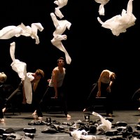 Dancers on a dark stage throw white scarves into the air. They wear black clothes and move in sync., © Regina Brocke