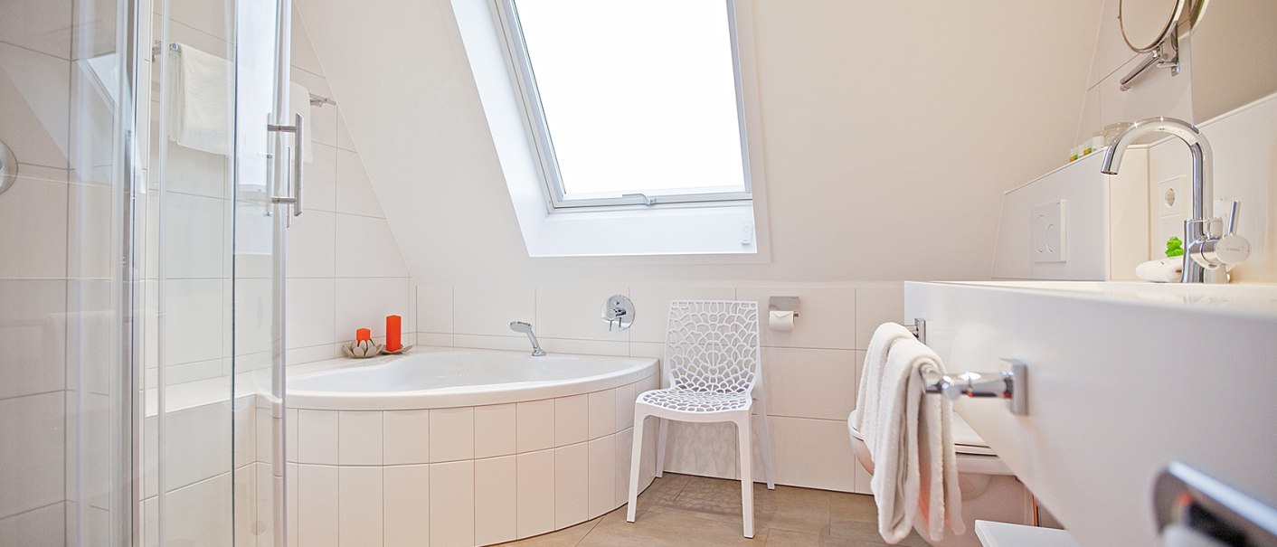 Helles Badezimmer mit Eckbadewanne, Glasdusche, Waschbecken und Dachfenster. Ein wei&szlig;er Stuhl und ein orangefarbener Teppich erg&auml;nzen das Design., &copy; Hotel Krone