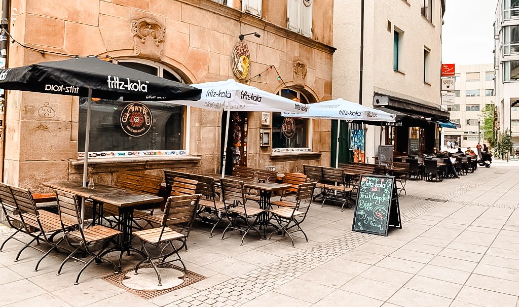 Ein Café mit Sitzgelegenheiten im Freien, Sonnenschirmen und einer Tafel mit Angeboten. Das Gebäude hat eine historische Fassade., © Mata Hari