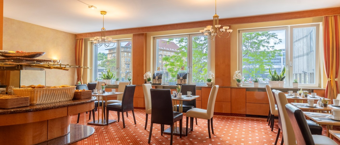 A bright breakfast room with laid tables, large windows and warm light. In the foreground are baskets of pastries., © PLAZA INN Rieker Stuttgart Hauptbahnhof
