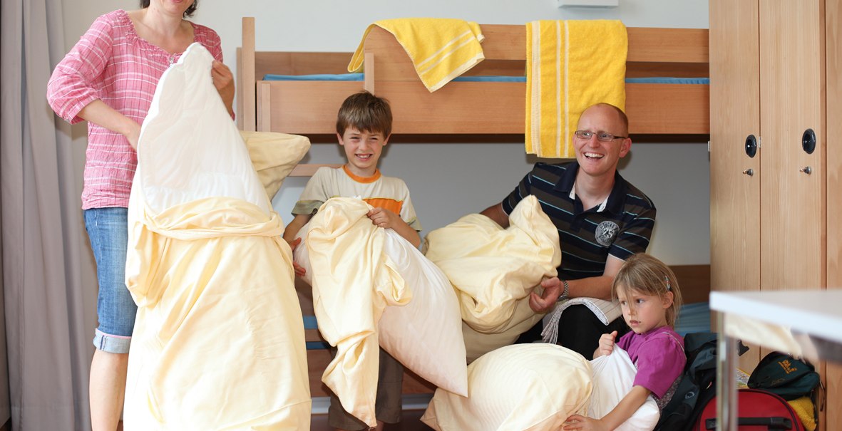 A family in a room with a bunk bed. They are holding yellow bedding and smiling. Towels are hanging on the bed. A child is sitting on a suitcase., © DJH Landesverband Baden-Württemberg e.V. A family in a room with a bunk bed. They are holding yellow bedding and smiling. Towels are hanging on the bed. A child is sitting on a suitcase., © DJH Landesverband Baden-Württemberg e.V.