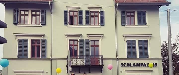 Three-storey building with 'Schlampazius' lettering, colorful balloons and people in front of the entrance. Historic architecture with red window frames., © Mora Fütterer Three-storey building with 'Schlampazius' lettering, colorful balloons and people in front of the entrance. Historic architecture with red window frames., © Mora Fütterer