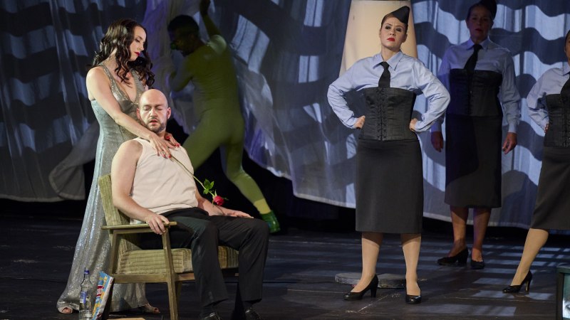 Theaterszene mit einer Frau in einem glitzernden Kleid, die einem sitzenden Mann eine Rose reicht. Weitere Personen in Uniformen stehen im Hintergrund., © Martin Sigmund Theaterszene mit einer Frau in einem glitzernden Kleid, die einem sitzenden Mann eine Rose reicht. Weitere Personen in Uniformen stehen im Hintergrund., © Martin Sigmund