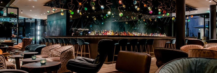 Moderne Bar mit stilvollen Sesseln und Tischen, bunte h&auml;ngende Lichter und eine lange Theke mit Barhockern., &copy; Motel One - Stuttgart Hauptbahnhof