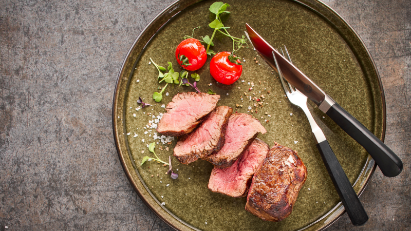 Ein Teller mit geschnittenem Steak, zwei Kirschtomaten und Besteck auf einem rustikalen Untergrund., © MAREDO Ein Teller mit geschnittenem Steak, zwei Kirschtomaten und Besteck auf einem rustikalen Untergrund., © MAREDO