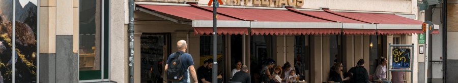 Ein Café mit rotem Vordach und dem Schriftzug 'Queer Kaffeehaus'. Menschen sitzen draußen an Tischen. Ein Mann geht vorbei., © SMG, Sarah Schmid