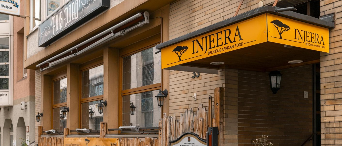 Restaurant Injeera mit gelbem Schild und Holzzaun, bietet afrikanische Küche an. Außenansicht mit Laternen und Pflanzen., © SMG, Sarah Schmid Restaurant Injeera mit gelbem Schild und Holzzaun, bietet afrikanische Küche an. Außenansicht mit Laternen und Pflanzen., © SMG, Sarah Schmid