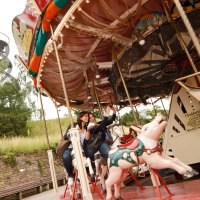 Zwei Personen fahren auf einem nostalgischen Karussell mit Pferdefiguren. Im Hintergrund ist ein spiralförmiger Aussichtsturm zu sehen., © SMG, Christoph Düpper Zwei Personen fahren auf einem nostalgischen Karussell mit Pferdefiguren. Im Hintergrund ist ein spiralförmiger Aussichtsturm zu sehen., © SMG, Christoph Düpper