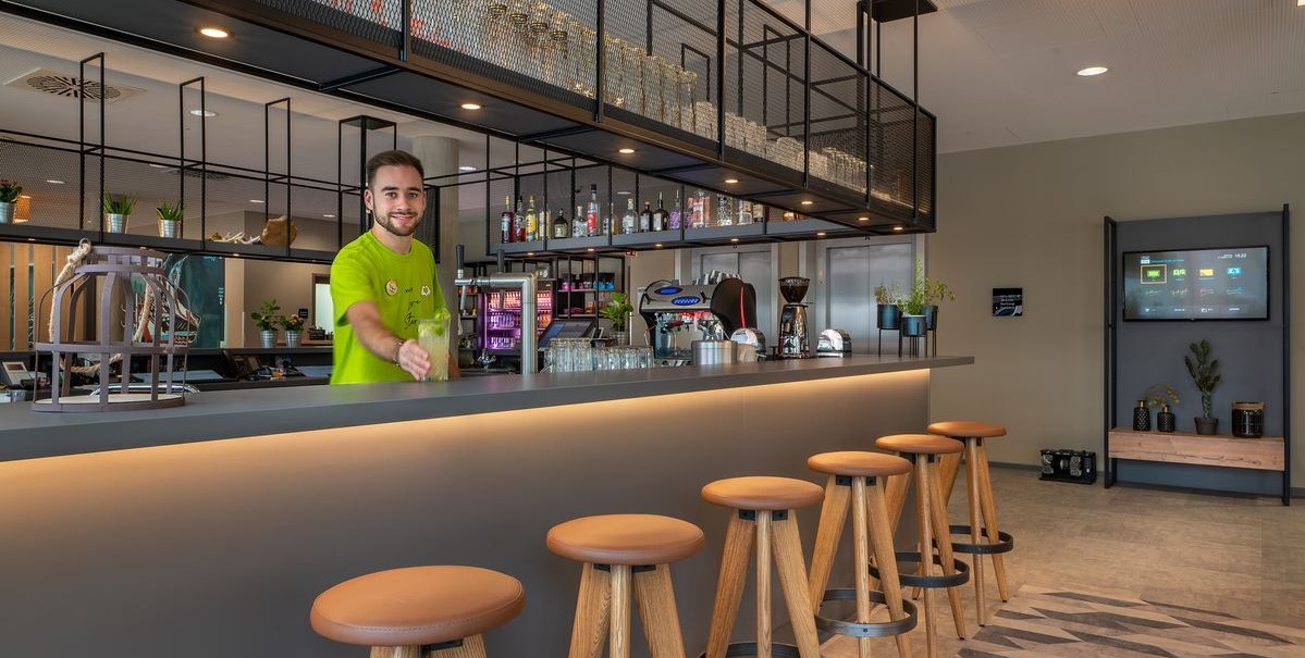 Ein Barkeeper in einem modernen Barbereich mit hohen Hockern, Regalen mit Gläsern und Flaschen, und einer gemütlichen Atmosphäre., © Novum Hospitality Ein Barkeeper in einem modernen Barbereich mit hohen Hockern, Regalen mit Gläsern und Flaschen, und einer gemütlichen Atmosphäre., © Novum Hospitality