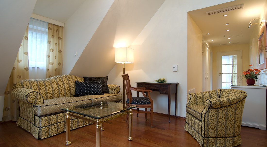 Eleganter Wohnbereich mit gemustertem Sofa, Sessel, Glastisch und Schreibtisch. Warme Beleuchtung und Holzboden schaffen eine gemütliche Atmosphäre., © Jeanette Bender