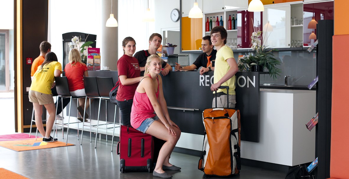 Young people with luggage at a modern hotel front desk. Some are sitting, others are standing while they check in. A bright and friendly atmosphere., © DJH Landesverband Baden-Württemberg e.V. Young people with luggage at a modern hotel front desk. Some are sitting, others are standing while they check in. A bright and friendly atmosphere., © DJH Landesverband Baden-Württemberg e.V.