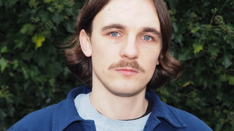 Ein Mann mit braunen Haaren und Schnurrbart steht vor einem Hintergrund aus grünen Blättern. Er trägt eine blaue Jacke und ein graues Shirt., © Jakob Kielgaß Ein Mann mit braunen Haaren und Schnurrbart steht vor einem Hintergrund aus grünen Blättern. Er trägt eine blaue Jacke und ein graues Shirt., © Jakob Kielgaß