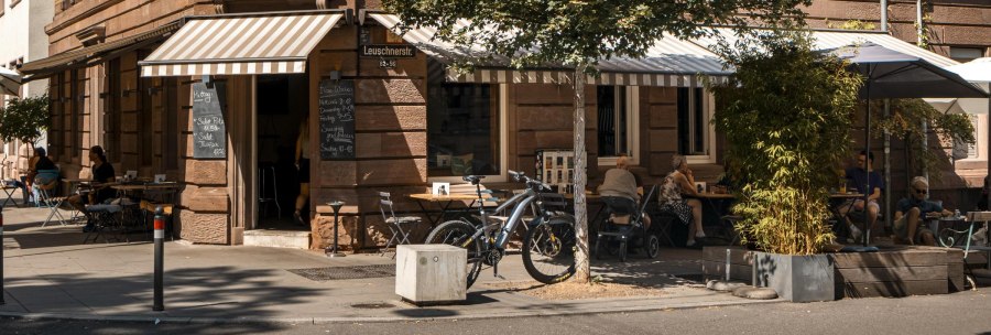 Ein gem&uuml;tliches Stra&szlig;encaf&eacute; mit gestreiften Markisen, Tischen und St&uuml;hlen im Freien. G&auml;ste sitzen im Schatten, Fahrr&auml;der sind abgestellt., &copy; SMG, Sarah Schmid