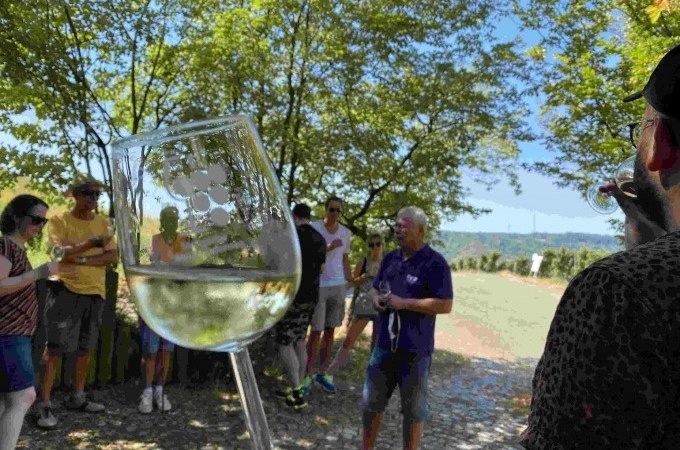 Gruppe bei einer Weinverkostung im Freien. Ein Glas Wei&szlig;wein wird in den Vordergrund gehalten. B&auml;ume und blauer Himmel im Hintergrund., &copy; Weingenuss Stuttgart