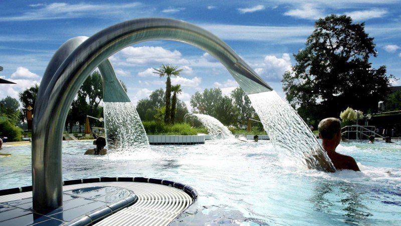 Ein Außenpool mit Wasserfontänen und Palmen, umgeben von Bäumen und einem Gebäude. Menschen genießen das Wasser unter blauem Himmel., © Stuttgarter Bäder Ein Außenpool mit Wasserfontänen und Palmen, umgeben von Bäumen und einem Gebäude. Menschen genießen das Wasser unter blauem Himmel., © Stuttgarter Bäder