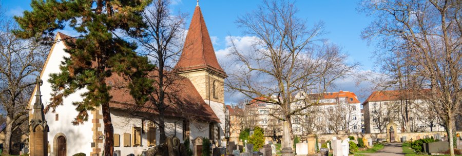 The Uff Cemetery, with its church, gravestones, and trees under a blue sky; residential buildings can be seen in the background., © Peter Jagusch/Landeshauptstadt Stuttgart The Uff Cemetery, with its church, gravestones, and trees under a blue sky; residential buildings can be seen in the background., © Peter Jagusch/Landeshauptstadt Stuttgart